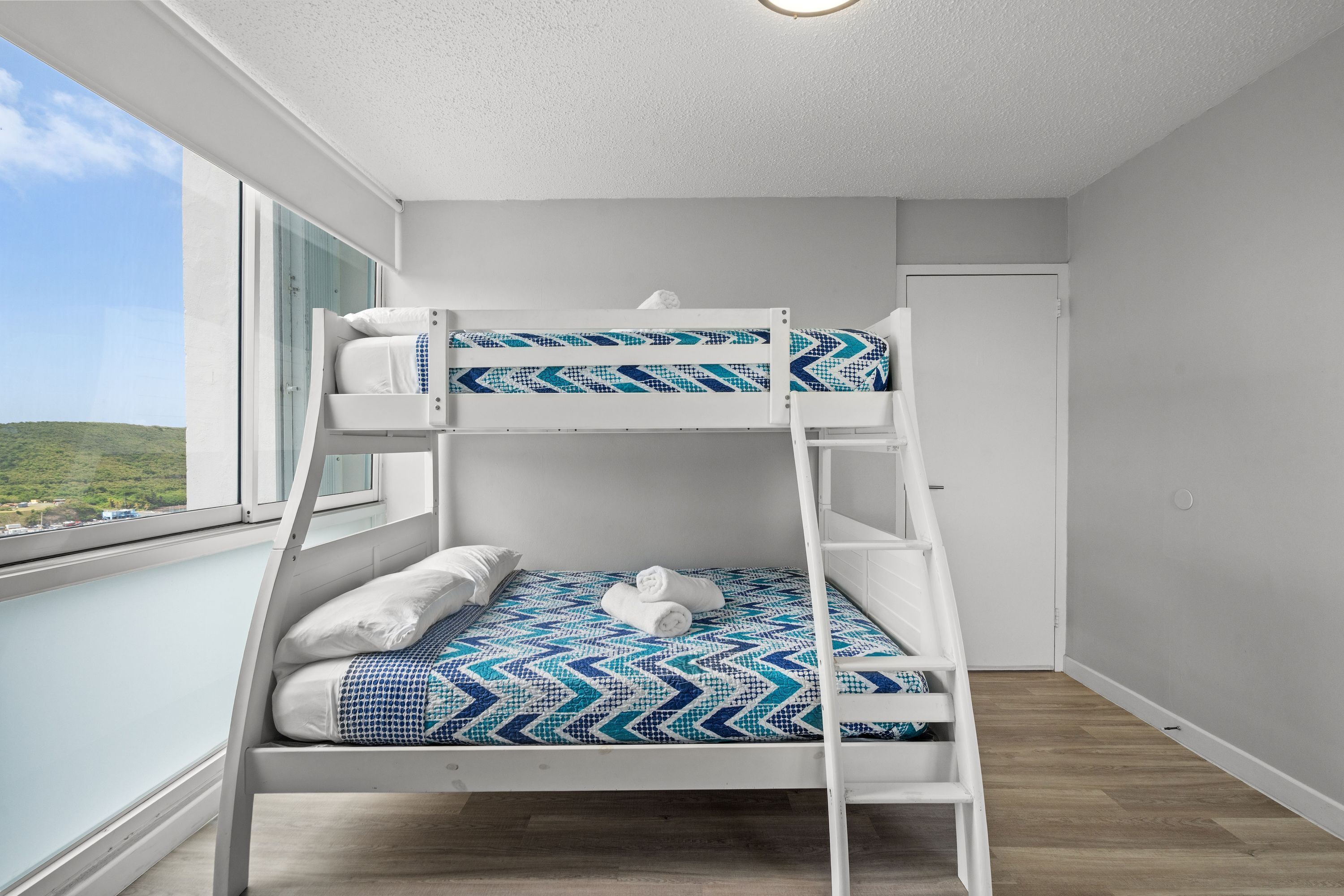A bunk bed with a patterned bedspread, a white headboard, and a view of the outdoors through a window.