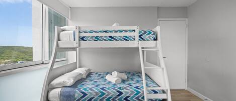 A bunk bed with a patterned bedspread, a white headboard, and a view of the outdoors through a window.