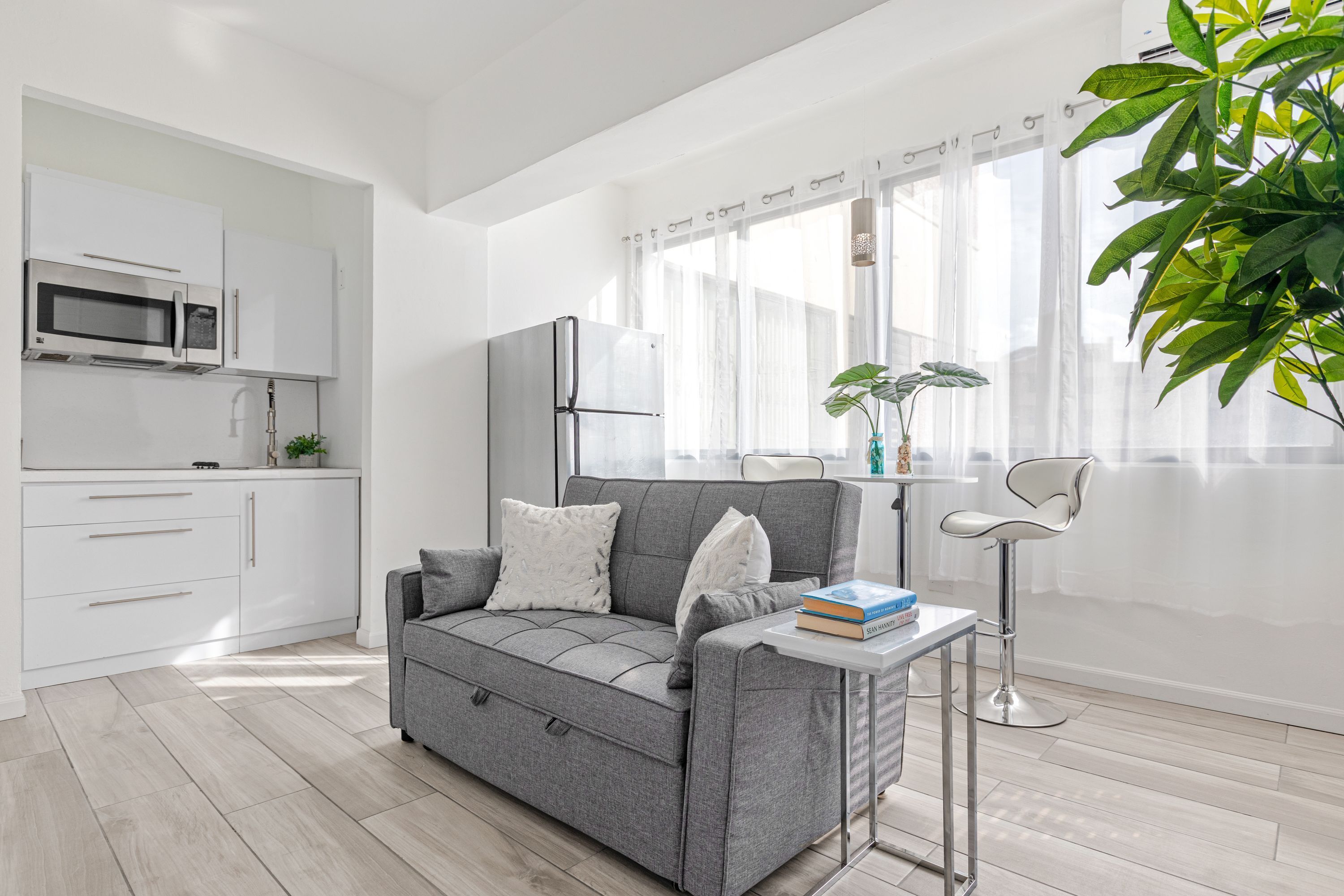 Eine moderne Küche mit weißen Schränken, einer Mikrowelle und einem Kühlschrank. Ein graues Sofa mit Kissen und ein kleiner Tisch mit einem Buch und einer Vase stehen dort. Im Eckbereich ist eine Pflanze zu sehen.
