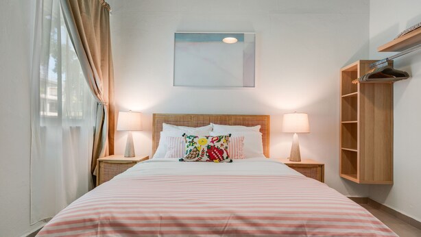 A neatly made bed with a striped bedspread, two bedside lamps, a wooden headboard, a wall-mounted shelf, and a window with curtains. - 2bl-piccioni in San Juan (San Juan)