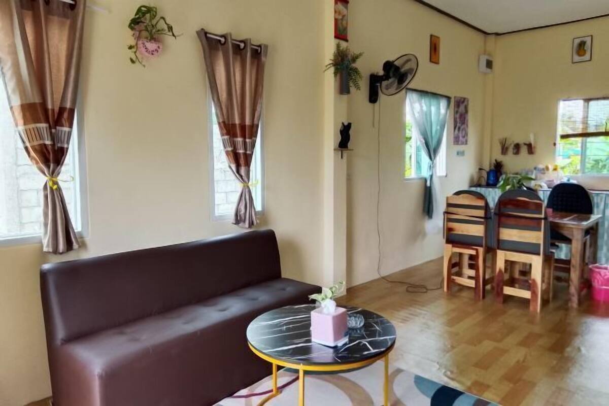 A room with a brown sofa, a round table with a marble top, wooden chairs, a fan, and a window with curtains.