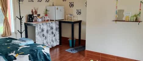 Una habitación con una cama, un escritorio, una silla, un refrigerador y una estantería con objetos.
