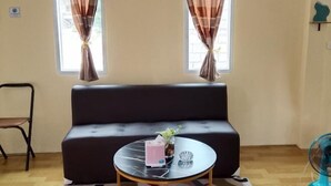 Ein Zimmer mit einem dunklen Sofa, einem runden Tisch mit einer Vase und einer Papprohhdose, sowie einem Standventilator.