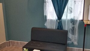 Ein kleines, dunkelgraues Sofa mit einer weißen Kiste darauf, vor einer türkisfarbenen Wand mit einem Fenster mit transparenten Vorhängen mit tropischen Blattmustern.
