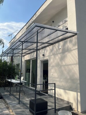 A modern outdoor patio with a glass enclosure, a table, chairs, and a black ottoman.