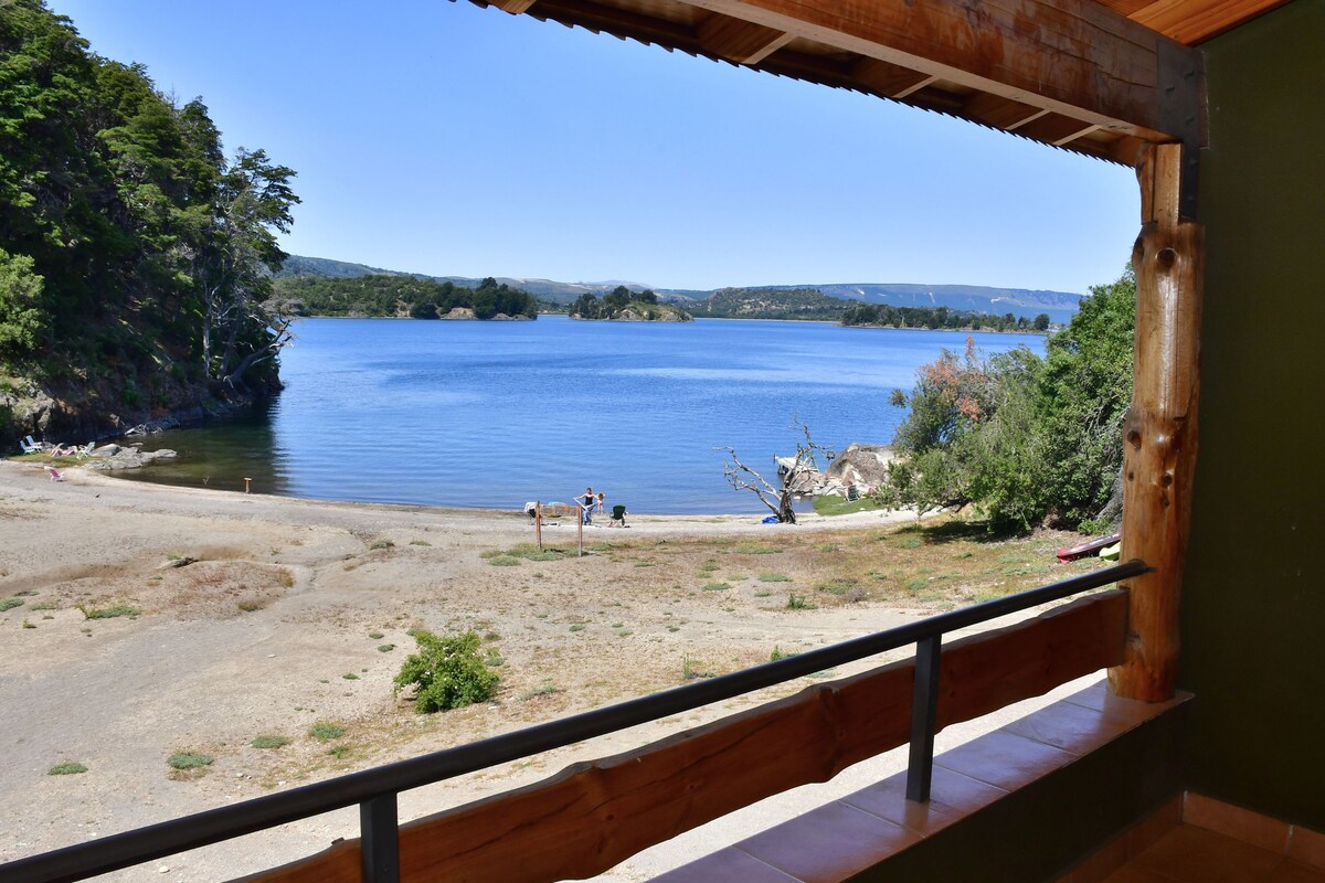 Blick auf einen See von einem Balkon mit Holzgeländer, ein Sandstrand und ein klarer, blauer Himmel.
