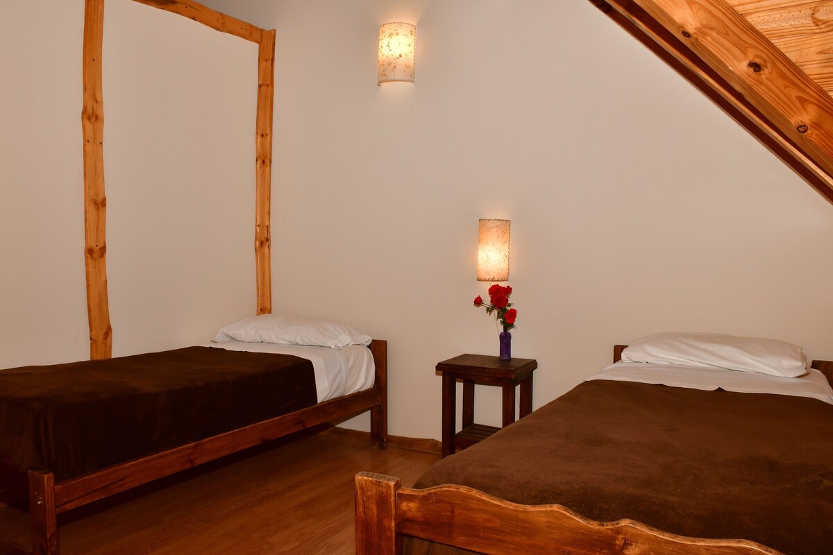 A room with two wooden beds, a small wooden nightstand with a vase of red flowers, and a wall-mounted light fixture.