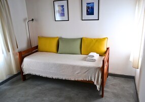 Una cama individual con un edredón blanco y dos almohadas, una amarilla y otra verde, en una habitación con una pared blanca lisa y un cuadro enmarcado sobre la cama.
