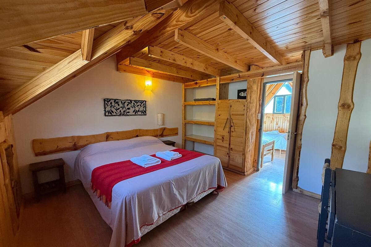 Un dormitorio de cabaña de madera con una cama, una mesita de noche, una lámpara de pared y una puerta de madera que da a un balcón.
