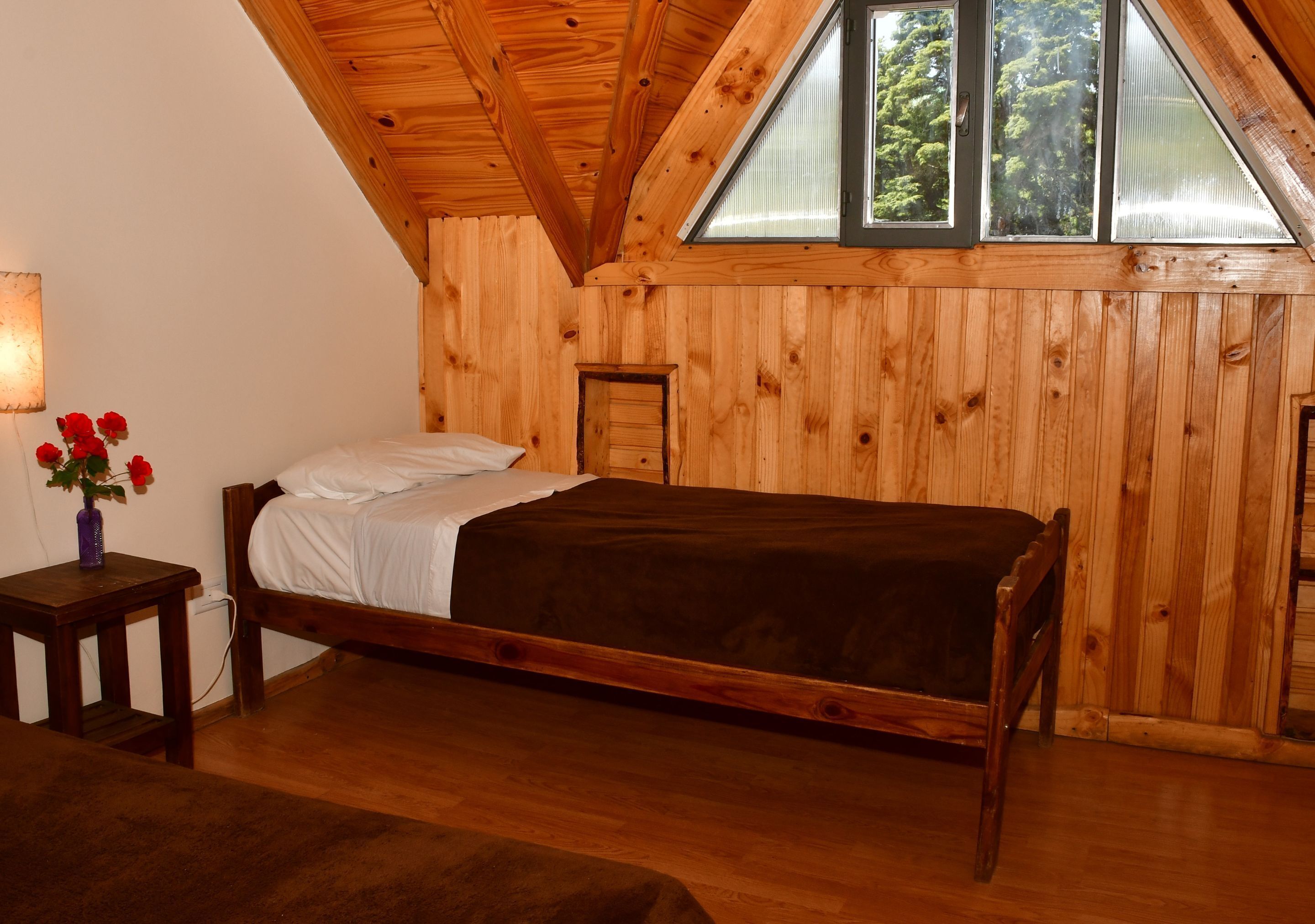 Una habitación de madera con una cama, una mesita con un florero y una ventana con vistas a árboles.