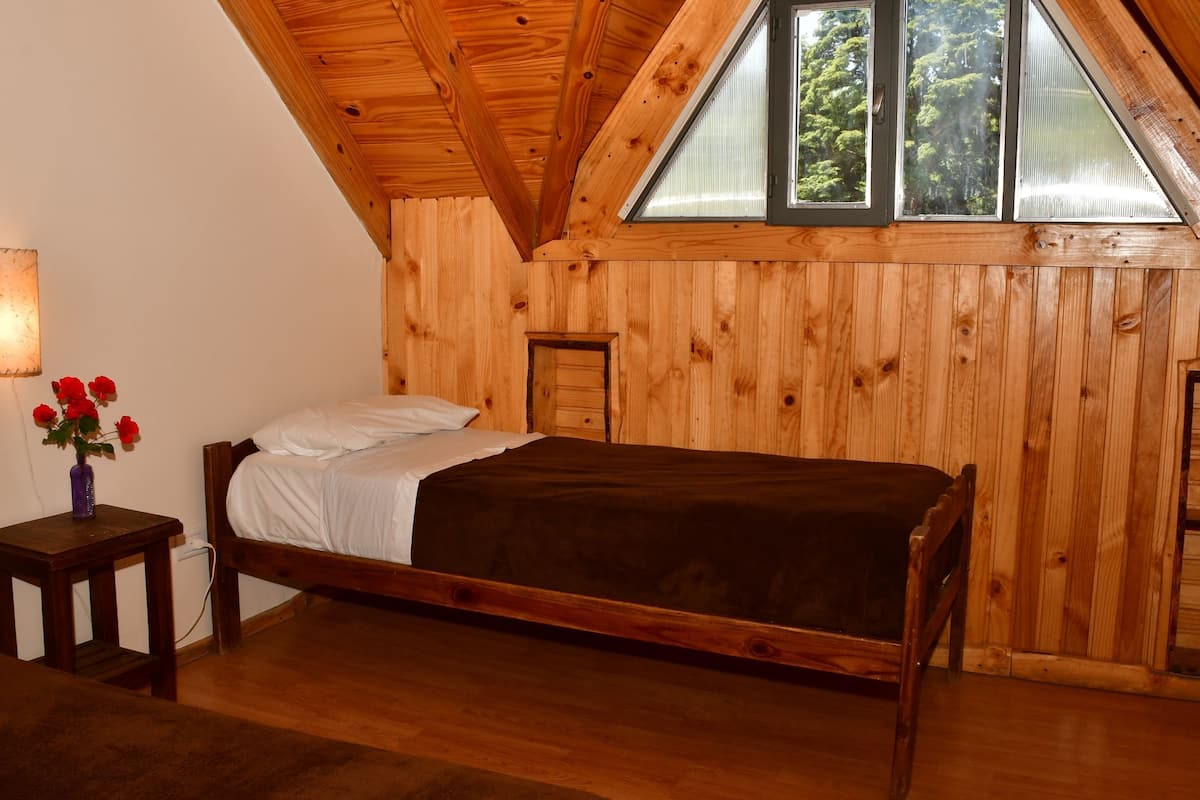 Una habitación de madera con una cama, una mesita con un florero y una ventana con vistas a árboles.