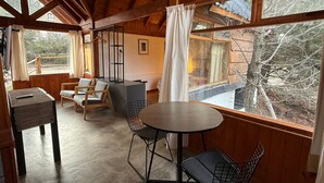 A cozy cabin interior with a wooden ceiling, a round table, two chairs, a small table with a remote control, and a view of the forest through large windows.