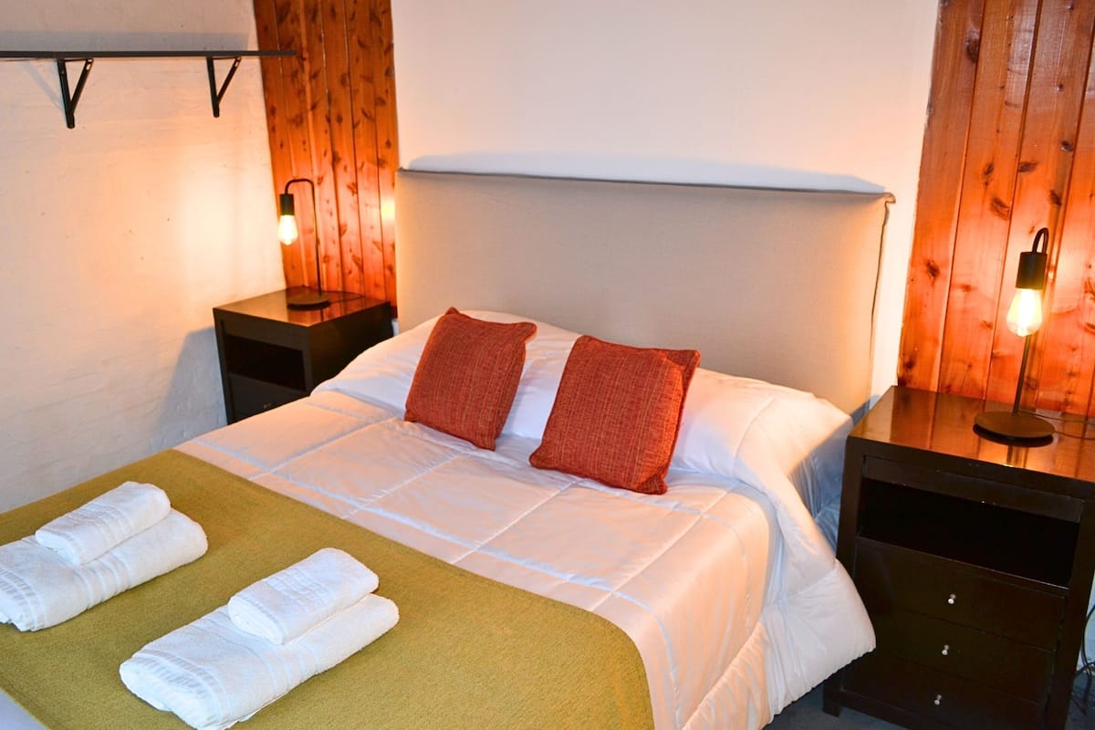 Una cama bien hecha con sábanas blancas, dos almohadas rojas y una manta verde. Hay dos mesitas de noche con lámparas, y las paredes tienen un diseño de paneles de madera.