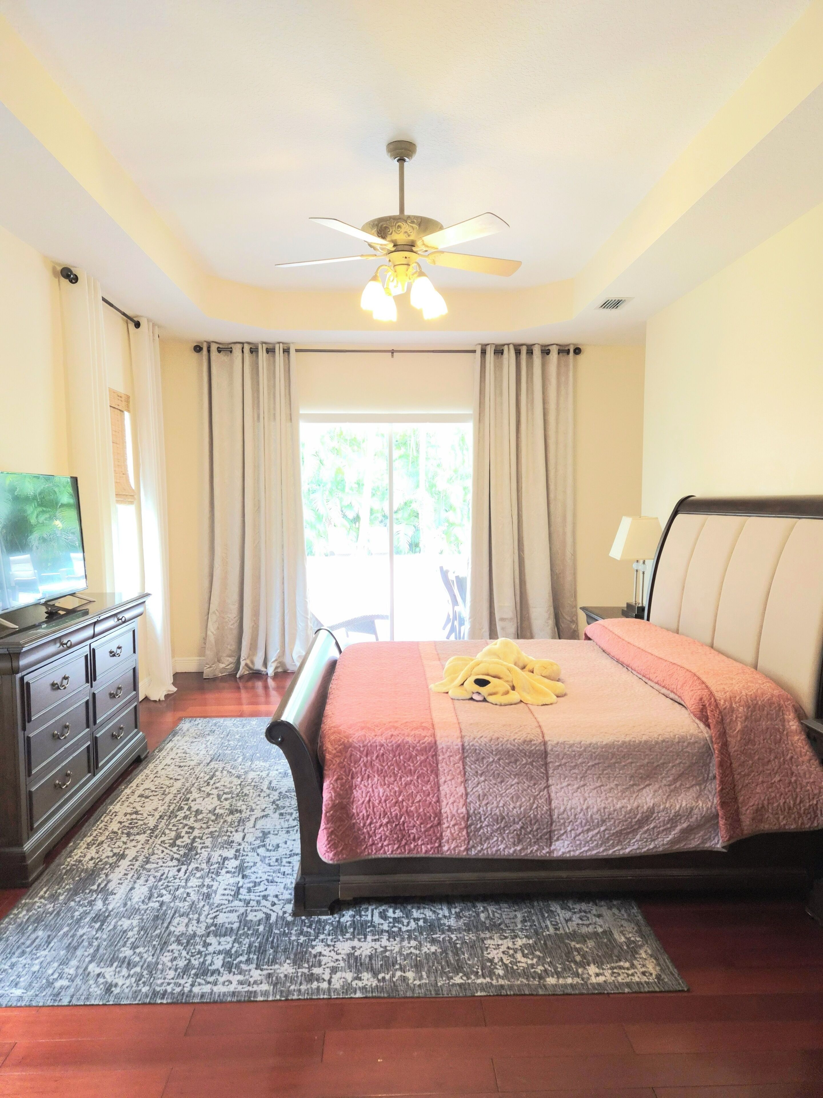 A bedroom with a bed, a ceiling fan, a television, and a window with curtains.