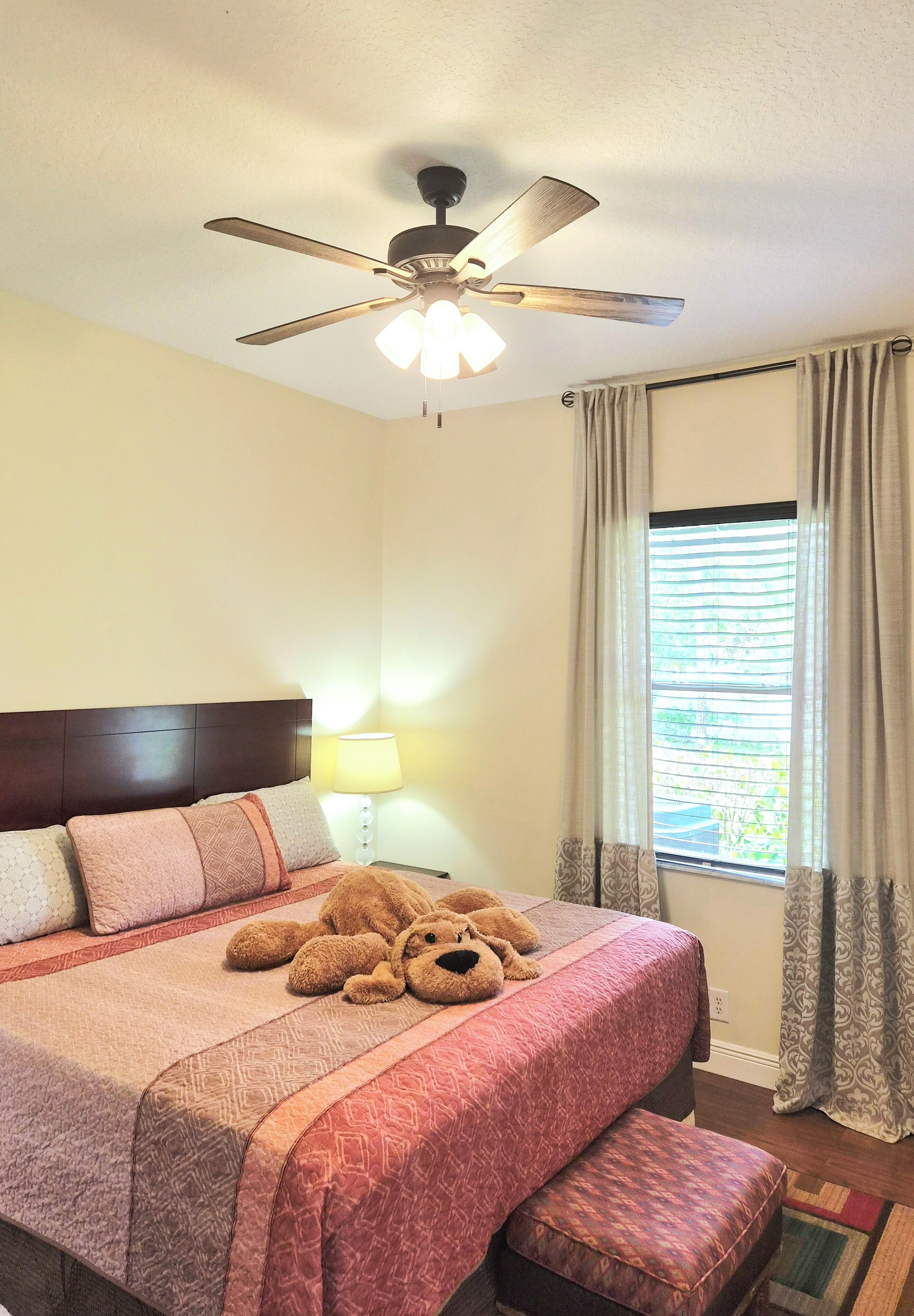 Une chambre d’hôtel avec un lit, un ventilateur de plafond, une fenêtre avec des rideaux et une table de chevet avec une lampe.