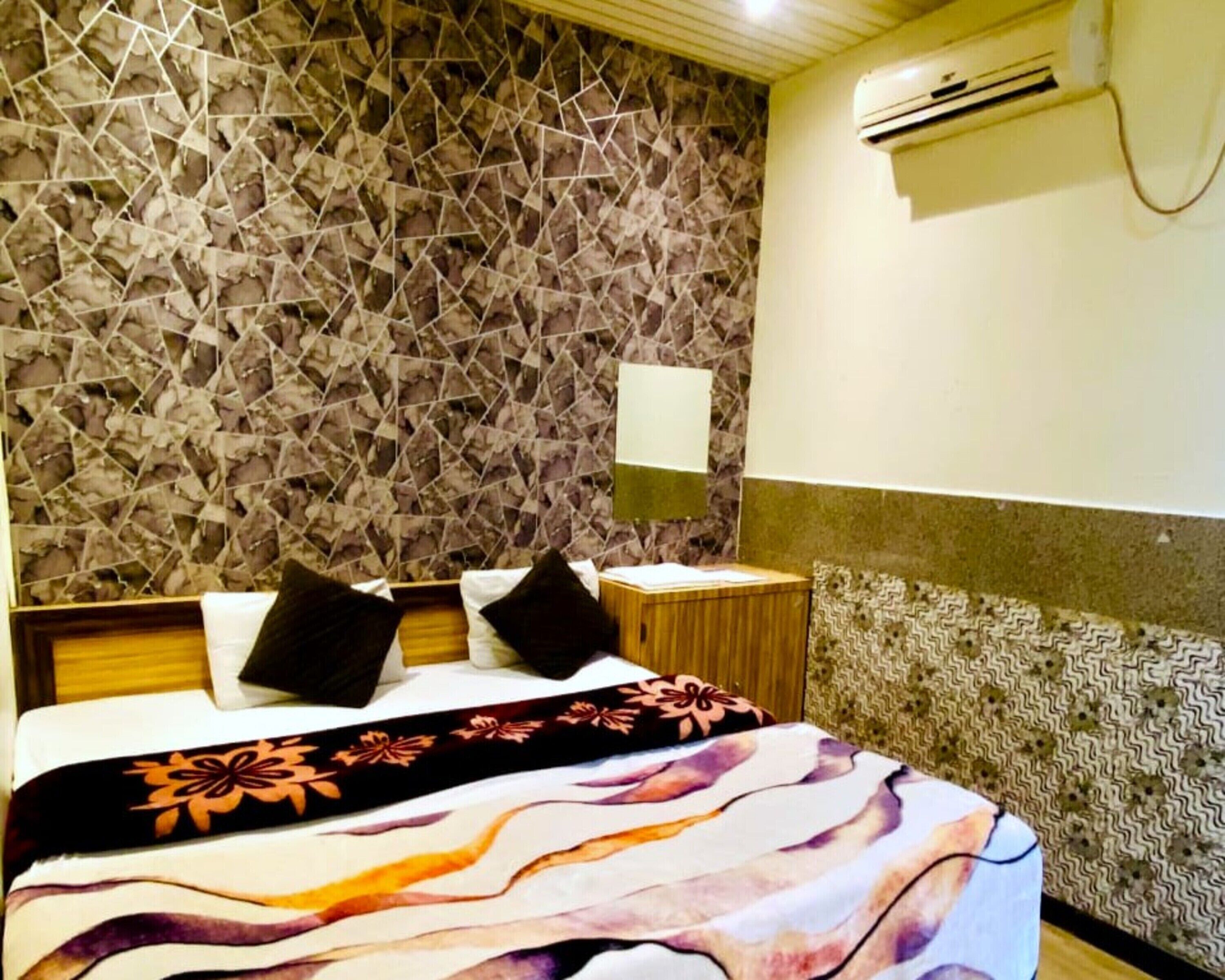 A hotel room with a patterned stone wall, a bed with a floral bedspread, a bedside table with a lamp, and an air conditioning unit on the wall.