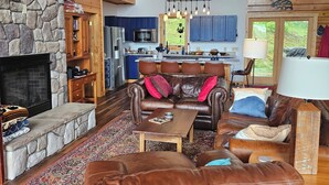A spacious living room with a stone fireplace, leather furniture, and a kitchen in the background. -  Beautiful 5-bedroom log cabin with spectacular mountain views. (Newland)