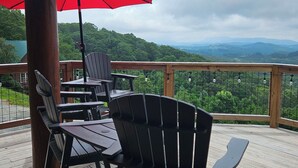 A wooden deck with outdoor seating, including chairs and tables, under a red umbrella, overlooking a mountainous landscape. -  Beautiful 5-bedroom log cabin with spectacular mountain views. (Newland)
