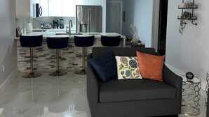 A modern kitchen with white cabinets, stainless steel appliances, and a central island. There are two bar stools and a dark grey sofa in the living area.