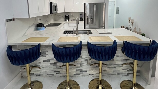 A modern kitchen with white cabinets, a stainless steel refrigerator, a built-in microwave, and a coffee maker. There are four blue bar stools with gold bases.