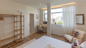 Un dormitorio moderno con cama, escritorio, silla, armario y un ventanal con vistas a edificios.