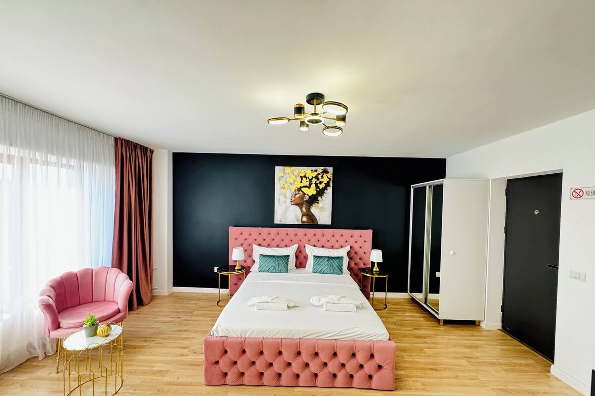 A modern bedroom with a pink tufted headboard, a large bed, a pink armchair, a small round table with a plant, and a painting of flowers on the wall.