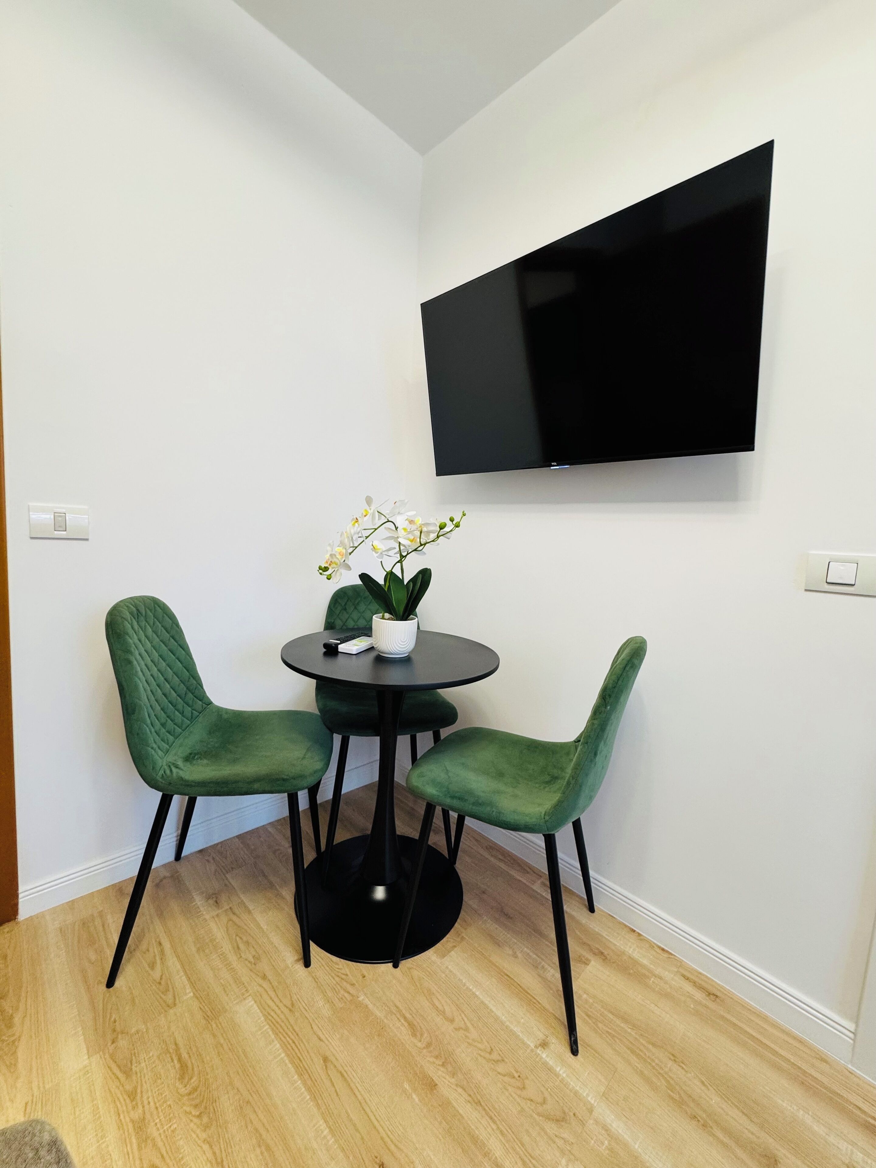 A small room with a round table, two green chairs, a white orchid plant, and a flat-screen TV mounted on the wall.