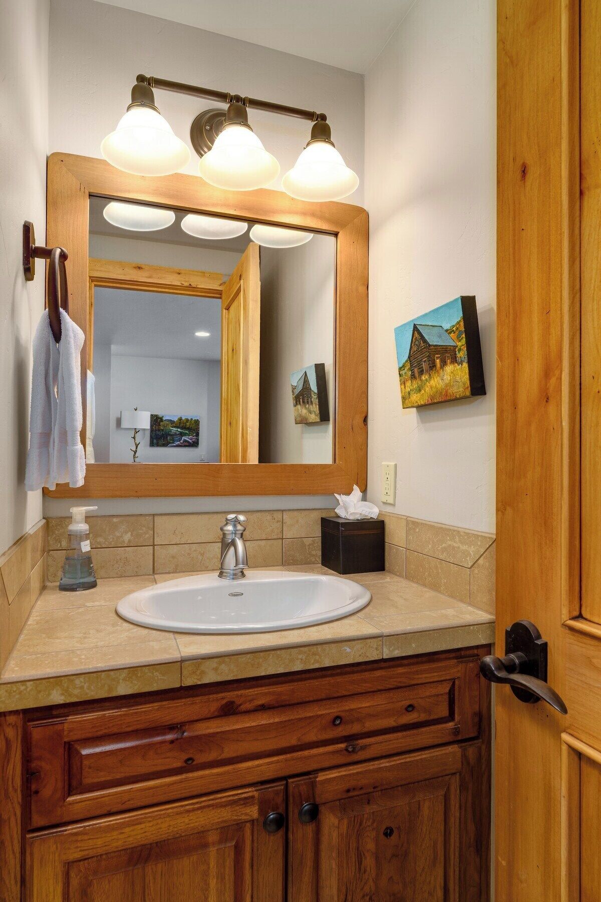 Un baño con tocador de madera, un lavabo blanco, un espejo con luces y un cuadro en la pared.