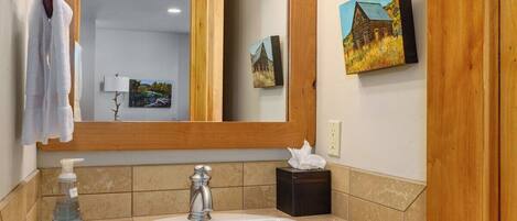 Un baño con tocador de madera, un lavabo blanco, un espejo con luces y un cuadro en la pared.