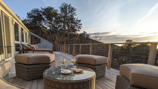 A deck with wicker furniture, a glass table, and two wine glasses. - Knockout Truro Retreat with Sweeping Views, Modern Amenities, Walk to Beach (Truro)