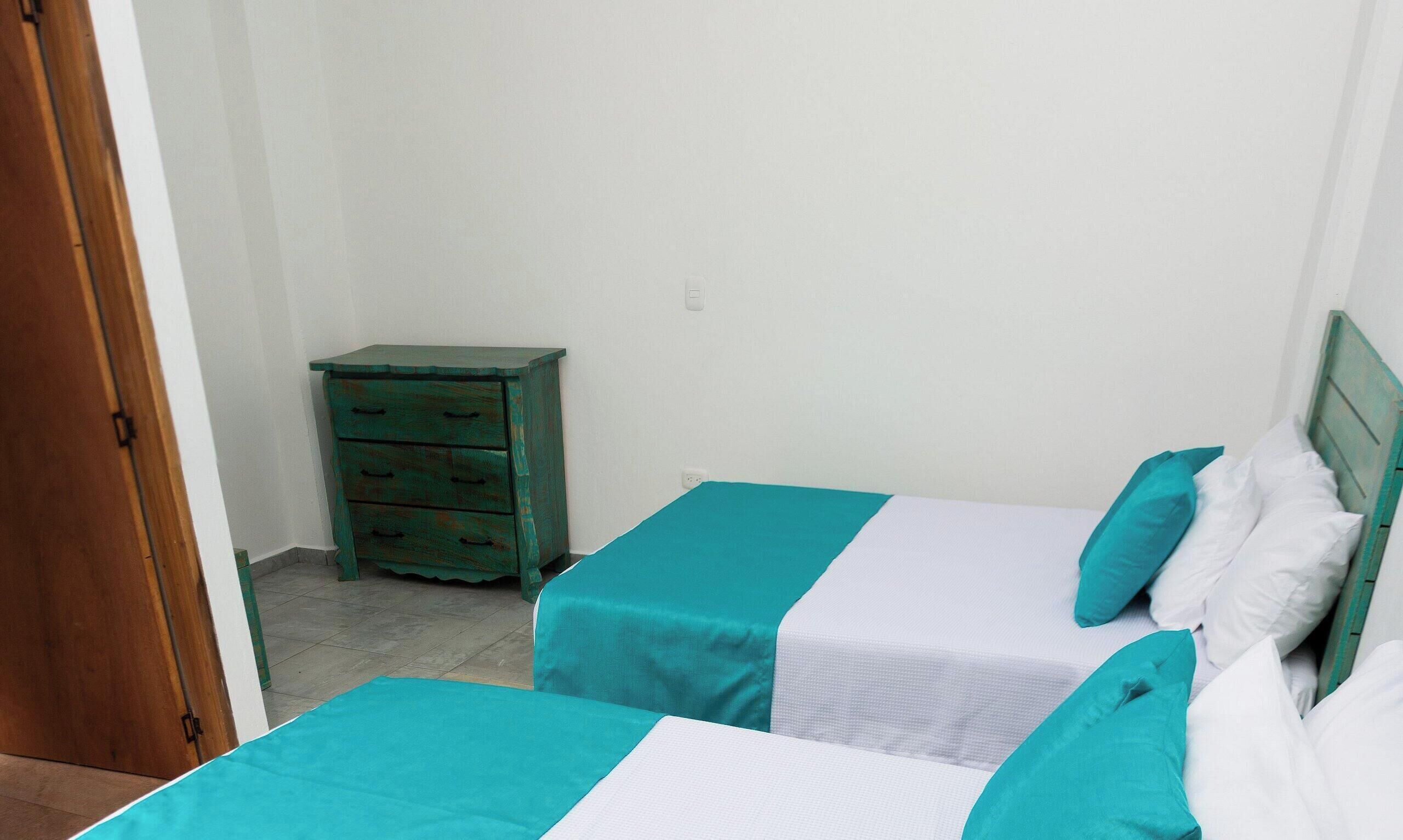 Dos camas individuales con ropa de cama verde azulada y blanca, una mesita de noche de madera y una pared blanca.