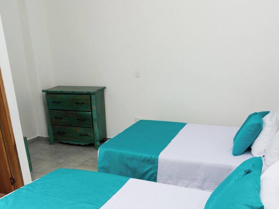 Dos camas individuales con ropa de cama verde azulada y blanca, una mesita de noche de madera y una pared blanca.