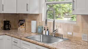 Una cucina con mobili bianchi, un piano di lavoro in granito, un lavello in acciaio inossidabile, una macchina per il caffè e una finestra vista verde.