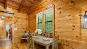 Ein Holzblockhaus-Interieur mit einem Esstisch für zwei Personen, einem Fenster mit Blick ins Grüne und einem kleinen, durch eine offene Tür einsehbaren Küchenbereich.