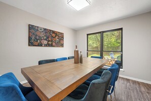 A conference room with a long wooden table, blue upholstered chairs, a large floral painting on the wall, and a window with a view of greenery. - Beautiful Spacious Home near OU Campus (Norman)