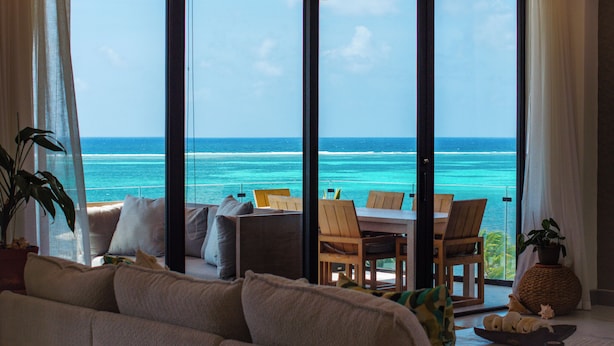 A room with a large sliding glass door leading to a balcony with a view of the ocean. There is a sofa, a dining table with chairs, and a potted plant. - Clearwater Belize (San Pedro)