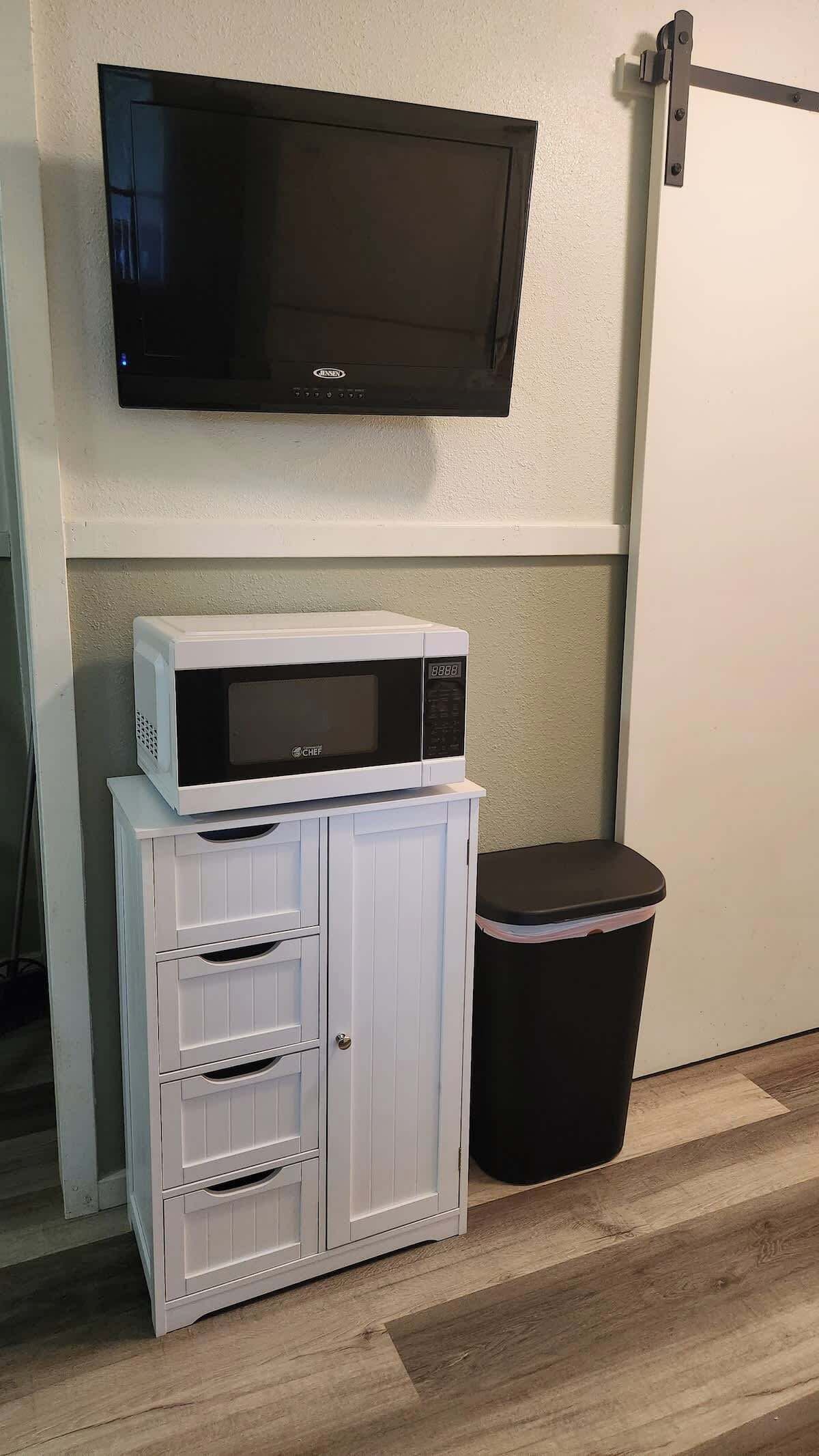 A white cabinet with drawers, a microwave, and a black trash bin against a wall.