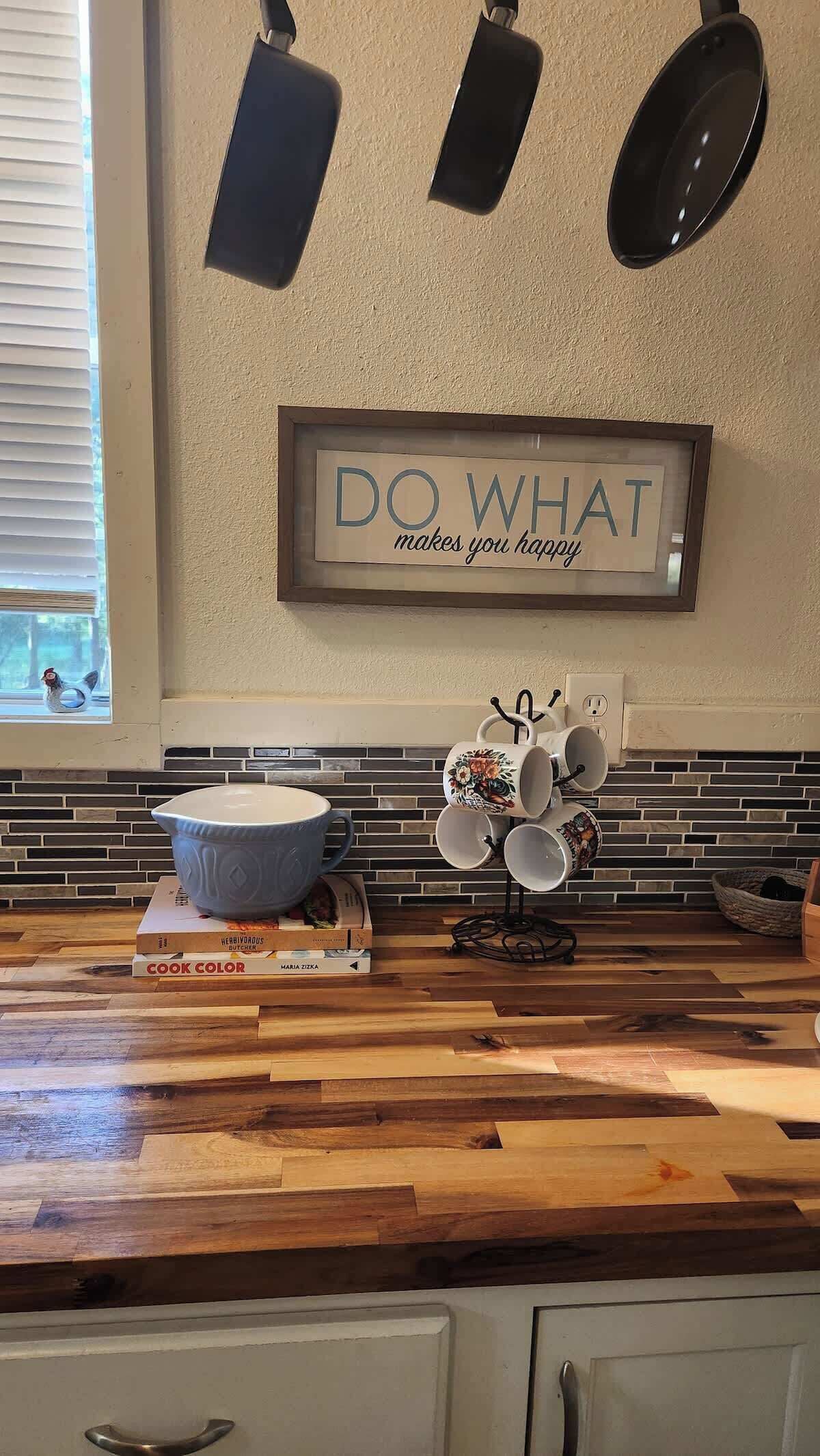 Una cocina con encimera de madera, un cartel enmarcado que dice 'DO WHAT makes you happy', y una ventana con persianas.