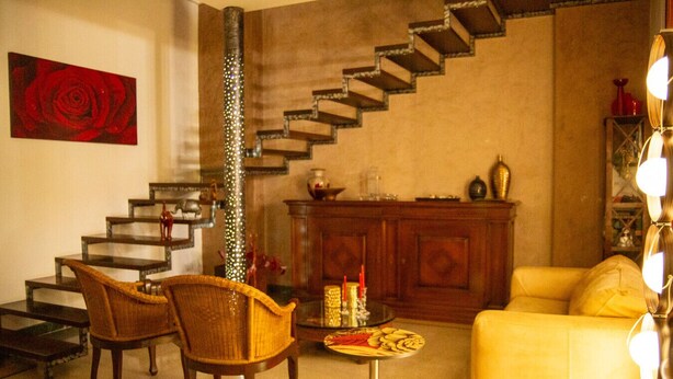 Ein Wohnzimmer mit Treppe, einem gelben Sofa, zwei Korbstühlen, einem kleinen runden Tisch mit Snacks und einem Gemälde einer roten Rose an der Wand.