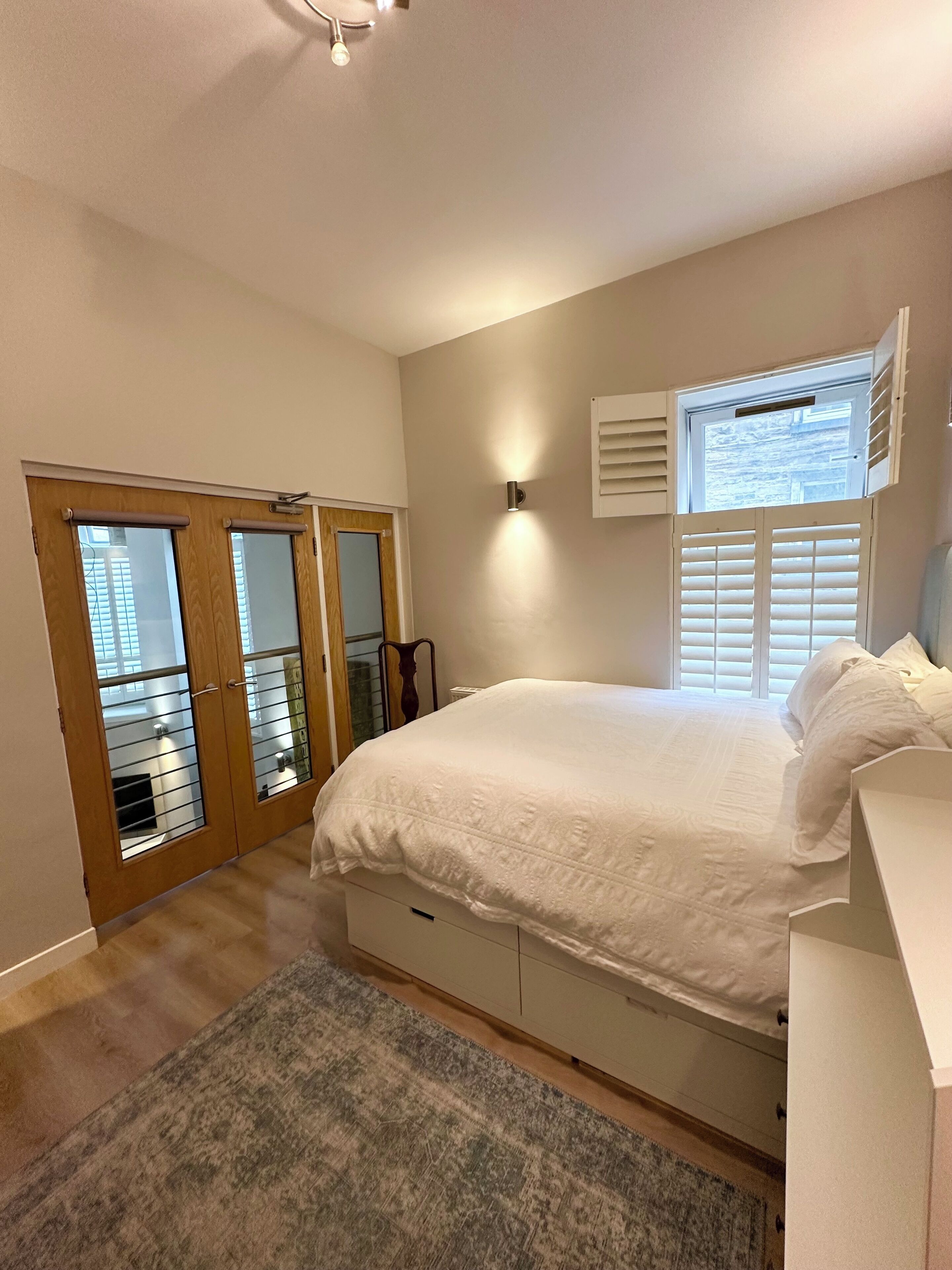 A bedroom with a bed, a dresser, a window with blinds, a door with a mirror, and a ceiling light.