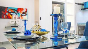 A dining table set with blue glassware, a white candle, and a colorful abstract painting on the wall. - Modern 1BR at Rizz Suites • GYM • Pool Access • Walk to Beach • King Bed• AC ew (Sosúa)