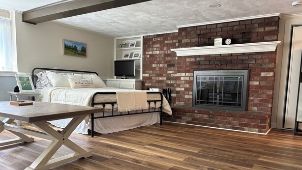 A modern bedroom with a brick fireplace, wooden flooring, a bed with white bedding, and a television on a white dresser.
