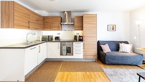 A compact kitchen with wooden cabinets, a built-in oven, and a microwave. There is a sofa with striped pillows in the adjacent living area.