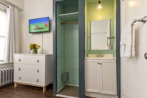 Une salle de bain avec un meuble-lavabo blanc, un miroir, une douche et un téléviseur fixé au mur.