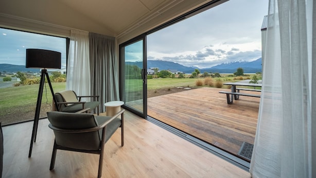 Una habitación moderna con un gran ventanal corredero que da a una terraza exterior. Adentro, hay un sofá, una pequeña mesa redonda y una lámpara de pie.
