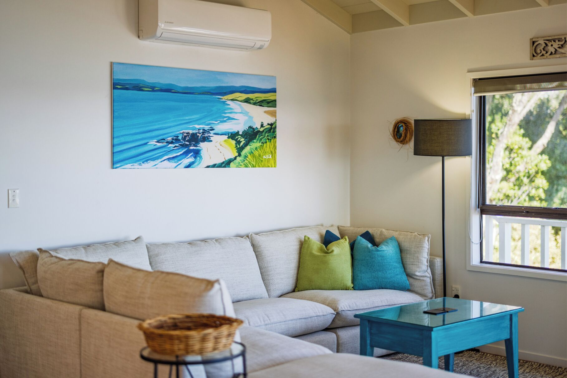 Un salon avec un canapé beige, une table basse en verre, un tableau représentant une plage et une fenêtre donnant sur un paysage verdoyant.