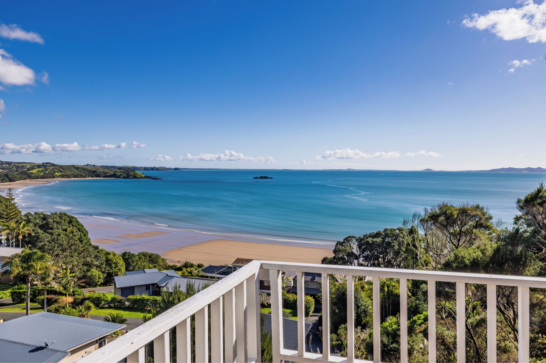 Une vue sur la côte, depuis un balcon donnant sur une plage aux eaux d’un bleu limpide, une île lointaine et une zone résidentielle avec des maisons et de la verdure.