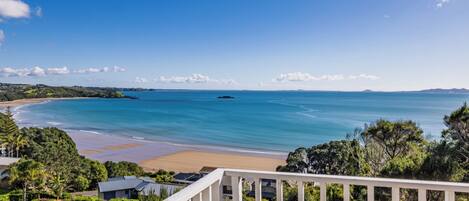 Une vue sur la côte, depuis un balcon donnant sur une plage aux eaux d’un bleu limpide, une île lointaine et une zone résidentielle avec des maisons et de la verdure.