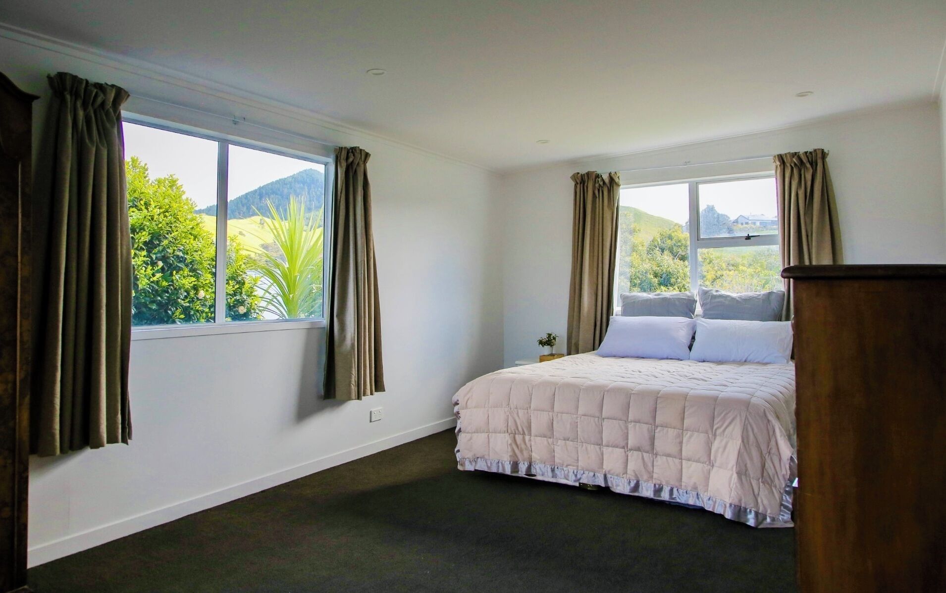 A bedroom with a bed, two windows with curtains, a view of greenery and hills, and a dark carpet.
