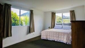 A bedroom with a bed, two windows with curtains, a view of greenery and hills, and a dark carpet.